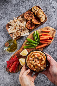 Draufsicht auf eine anonyme Person, die eine Schale mit leckerem Hummus neben eine Holzplatte mit verschiedenen Gemüsescheiben und Zitrone und ein Tablett mit Tortilla-Chips und Croutons auf einen grauen Marmortisch mit einer Flasche Olivenöl stellt - ADSF43084