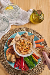 Draufsicht auf eine anonyme Person, die knusprige Tortilla-Chips in eine Schüssel mit leckerem Hummus eintaucht, umgeben von Gemüse und Croutons in der Nähe einer Flasche Olivenöl und Gläsern mit kalter Limonade auf einem Holztisch zu Hause - ADSF43085