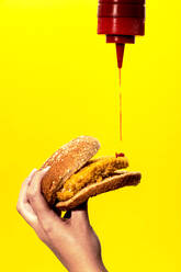 Crop anonyme weibliche Gießen Ketchup aus der Flasche auf frittierte Patty beim Essen leckeren Burger gegen gelben Hintergrund - ADSF43087