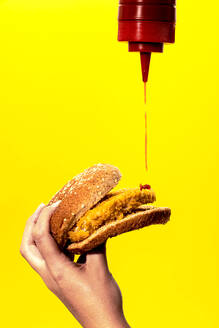 Crop anonyme weibliche Gießen Ketchup aus der Flasche auf frittierte Patty beim Essen leckeren Burger gegen gelben Hintergrund - ADSF43087