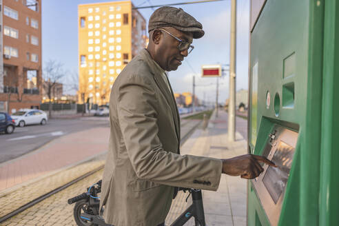 Älterer Geschäftsmann mit Elektrofahrzeug, der einen Geldautomaten auf dem Gehweg benutzt - JCCMF09255