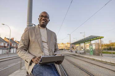 Smiling mature businessman with tablet PC by tramway - JCCMF09263