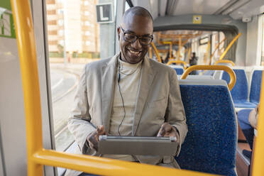 Lächelnder Geschäftsmann mit In-Ear-Kopfhörern und Tablet-PC in der Straßenbahn - JCCMF09266