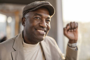 Smiling businessman with cap by window traveling in tram - JCCMF09274