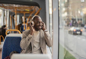 Glücklicher Pendler mit drahtlosen Kopfhörern und Blick aus dem Fenster in der Straßenbahn - JCCMF09276