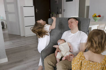 Vater mit kleinem Jungen entspannt sich auf einem Stuhl, während die Töchter zu Hause spielen - VIVF00355