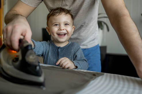 Glücklicher süßer Sohn hilft Vater beim Bügeln von Kleidung zu Hause - ANAF00968