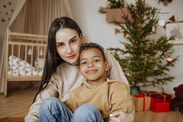 Lächelnde Mutter und Sohn sitzen zu Hause vor dem Weihnachtsbaum - MDOF00599