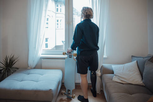 Man with skateboard looking through window at home - JOSEF16504