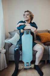 Happy young man with skateboard sitting on sofa at home - JOSEF16506