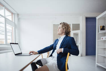 Thoughtful businessman sitting with laptop in office - JOSEF16513