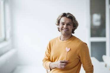 Happy man with heart shaped prop standing at home - JOSEF16537