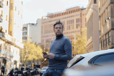 Nachdenklicher Mann mit Mobiltelefon, der sich in der Stadt an ein Auto lehnt - JOSEF16609