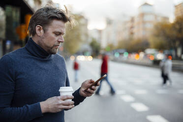 Mann mit Einweg-Kaffeebecher benutzt Mobiltelefon auf der Straße - JOSEF16635