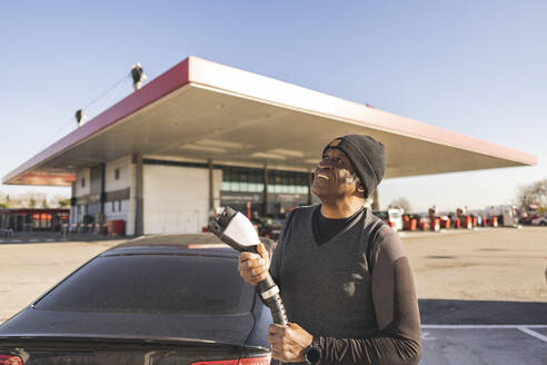 Smiling senior man holding power cord at charging station - JCCMF09304