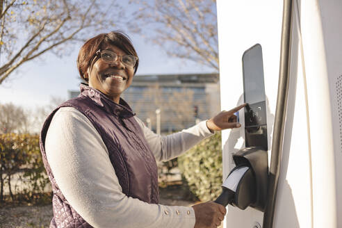 Smiling thoughtful senior woman standing by electric charging station - JCCMF09305