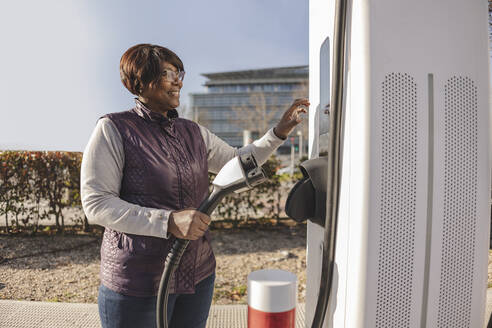 Woman standing with power cord at electric charging station - JCCMF09306