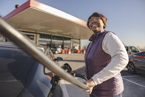 Happy senior woman charging electric car on sunny day - JCCMF09308