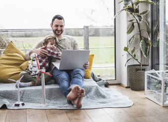Father and son looking at laptop sitting on sofa with wind turbines - UUF28065