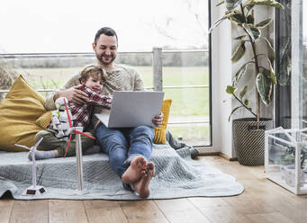 Vater und Sohn schauen auf einen Laptop und sitzen auf einem Sofa mit Windrädern - UUF28065