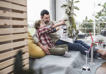 Father and son sitting on sofa playing with toy robot - UUF28067