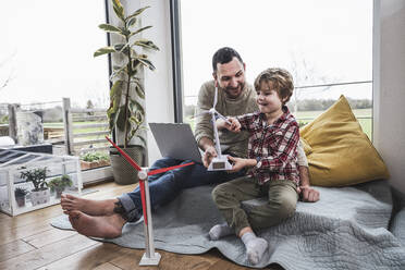 Father and son playing with wind turbine model sitting at home - UUF28069