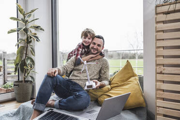 Happy father holding wind turbine model with son embracing from behind at home - UUF28075