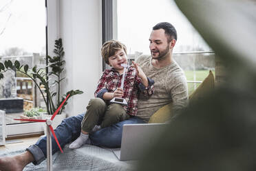 Son playing with wind turbine model sitting on father's lap at home - UUF28078