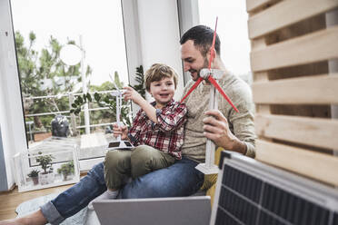 Smiling boy sitting with father holding wind turbine model sitting at home - UUF28080