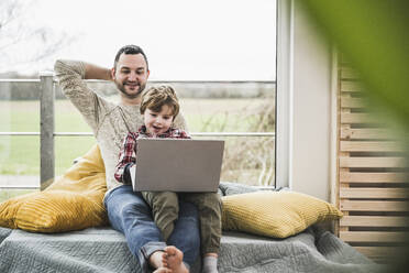 Lächelnder Vater und Sohn, die zu Hause gemeinsam Laptop schauen - UUF28091