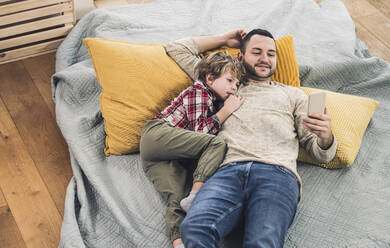 Lächelnder Vater, der ein Smartphone benutzt, während sein Sohn zu Hause auf einer Decke liegt - UUF28111