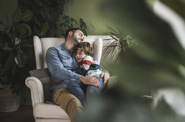 Lächelnder Vater entspannt sich mit seinem Sohn auf einem Sessel zu Hause - UUF28138