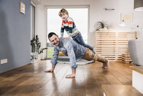 Boy balancing on back of father doing push-ups in living room at home - UUF28141