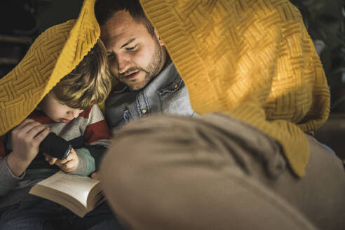 Vater und Sohn mit Taschenlampe lesen ein Buch unter einer gelben Decke - UUF28146