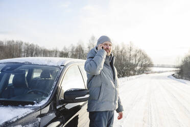 Lächelnder Mann in warmer Kleidung, der vor einem Auto auf einer winterlichen Straße mit seinem Smartphone telefoniert - EYAF02561