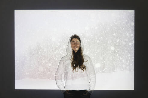 Happy woman with eyes closed standing in front of snowfall on projection screen - MRAF00916