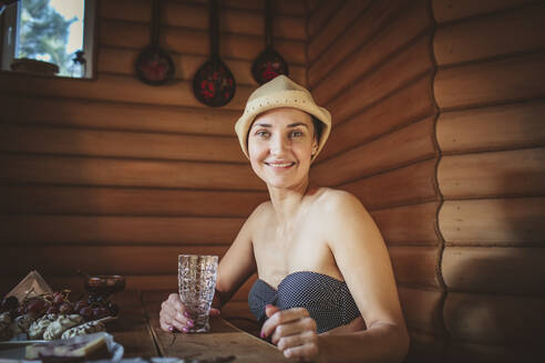 Lächelnde Frau mit Trinkglas am Tisch in der Sauna sitzend - MDOF00604