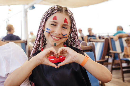 Lächelndes Mädchen mit Gesichtsbemalung gestikuliert Herzform auf Jacht - OSF01365