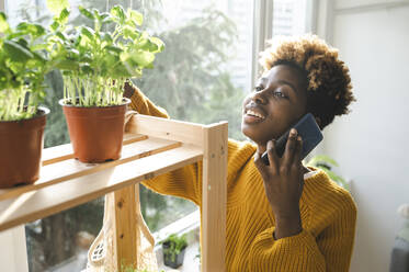 Glückliche Frau, die mit einem Smartphone telefoniert, mit Pflanzen auf einem Regal - ALKF00077