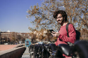 Glücklicher Mann, der eine mobile App nutzt, um an einem sonnigen Tag einen Elektroroller zu mieten - EBBF07823