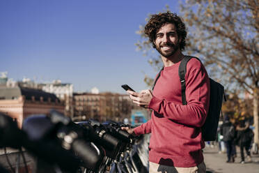 Smiling young man with smart phone standing at electric bicycle sharing system - EBBF07824