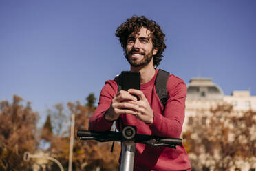 Smiling young man leaning on electric push scooter with smart phone - EBBF07833