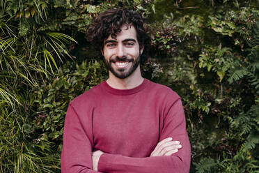 Happy man with arms crossed standing in front of plants - EBBF07839