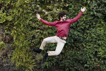 Carefree young man jumping in front of plants - EBBF07840