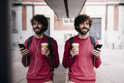 Glücklicher Mann mit Smartphone und Kaffeetasse auf dem Fußweg - EBBF07864