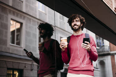 Smiling young man with smart phone and coffee cup standing at footpath - EBBF07865