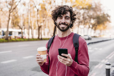 Fröhlicher junger Mann mit Mobiltelefon und Kaffeetasse auf dem Fußweg - EBBF07867