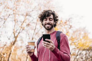 Glücklicher Mann, der ein Mobiltelefon benutzt und eine Kaffeetasse in der Hand hält, steht auf einem Fußweg - EBBF07869