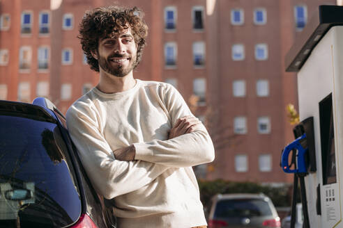 Lächelnder junger Mann, der an einem sonnigen Tag neben einem Elektroauto steht - EBBF07874