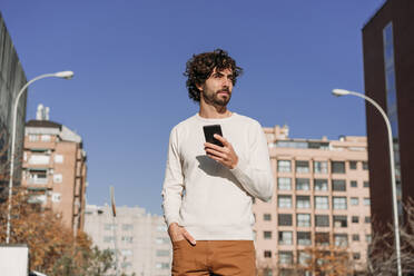 Thoughtful man with smart phone standing on sunny day - EBBF07875